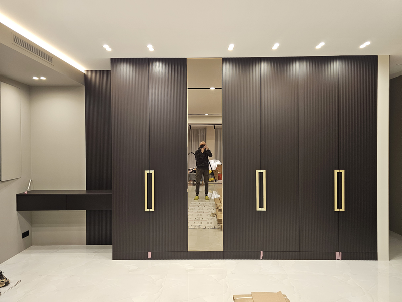 Корпусный шкаф Корпусный шкаф во всю стену с вертикально рифлёными фасадами тёмного оттенка, интегрированными ручками в отделке под латунь и центральной зеркальной дверью в полный рост, выполненный как архитектурный элемент интерьера с точной подгонкой по потолку и полу