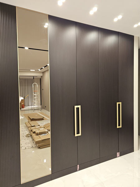 Фрагмент фасадной Фрагмент фасадной части корпусного шкафа с глубоким вертикальным рифлением, акцентными металлическими ручками прямоугольной формы и встроенной зеркальной секцией, подчёркивающей масштаб и геометрию пространства