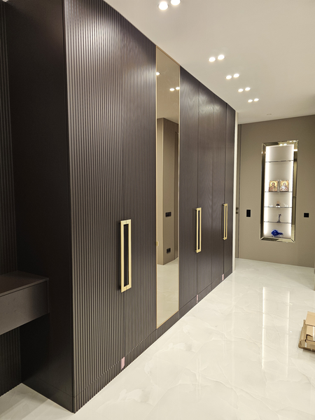 Корпусный шкаф Корпусный шкаф во всю стену с рифлёными распашными фасадами, зеркальной центральной дверью и акцентными вертикальными ручками, выполненный по индивидуальному проекту с точной геометрией и встроенным освещением