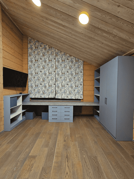 Общий вид Общий вид детской комнаты на мансарде с индивидуальной мебелью: длинная рабочая столешница вдоль стены, система хранения с ящиками и шкаф для одежды, спроектированные по размерам помещения и с учётом сложной геометрии.