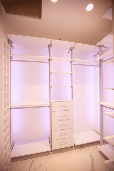 Светлая открытая гардеробная система с металлическими стойками, полками и встроенным высоким комодом, с акцентной подсветкой задних панелей, создающей эффект визуальной глубины и чистой архитектурной композиции.